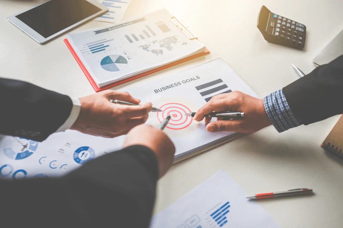 Two people are engaged in a business meeting, focusing on a document labeled "BUSINESS GOALS" and a target icon (bullseye symbol) on a desk.