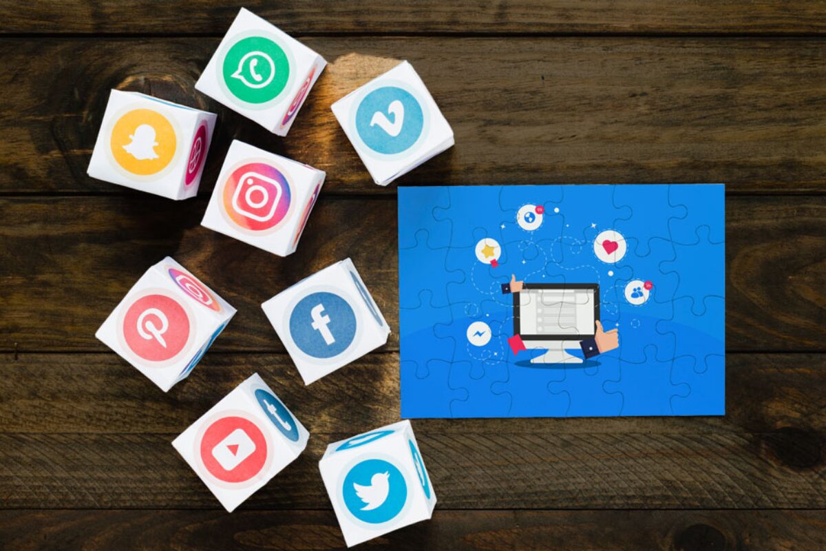 A collection of white cubes displaying various social media logos including WhatsApp, Snapchat, Instagram, Facebook, Twitter, and YouTube, alongside a blue jigsaw puzzle piece featuring a cartoon illustration of social media marketing and engagement on a computer screen.