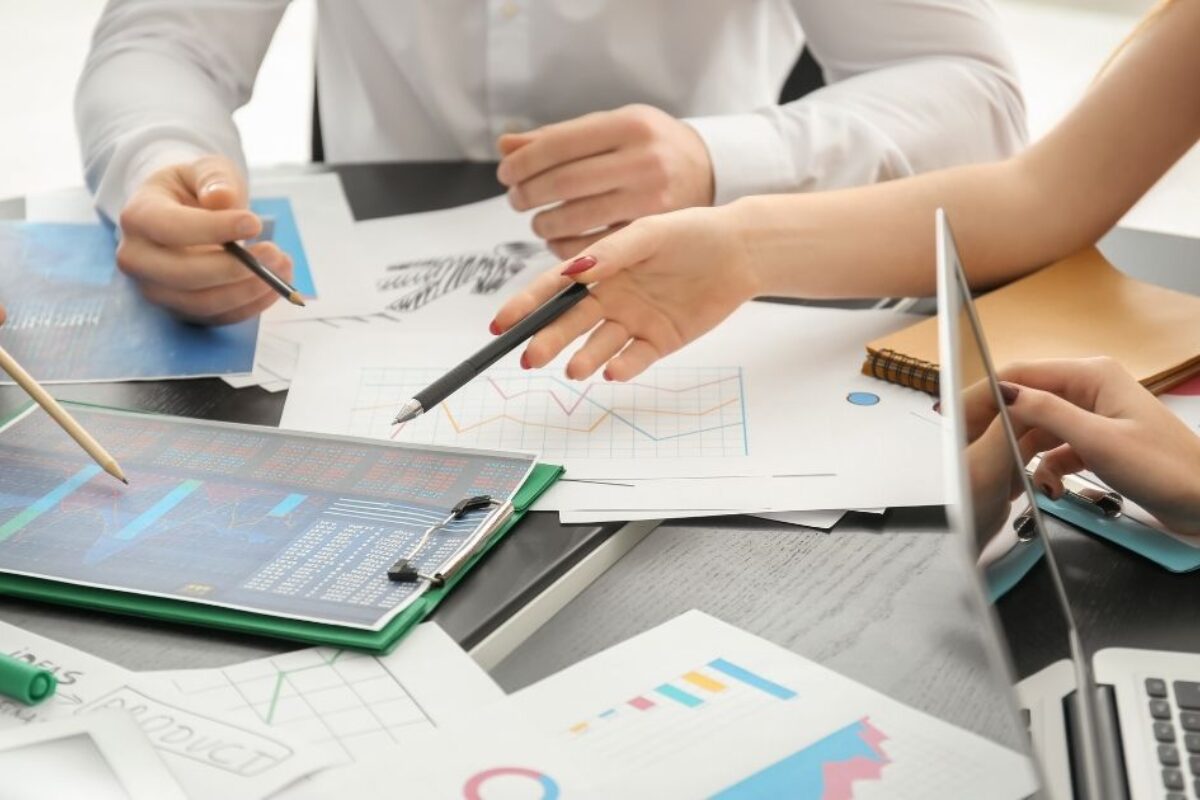 Two people in a business meeting are pointing at financial reports and line graphs on a desk filled with papers, a laptop, and a tablet.