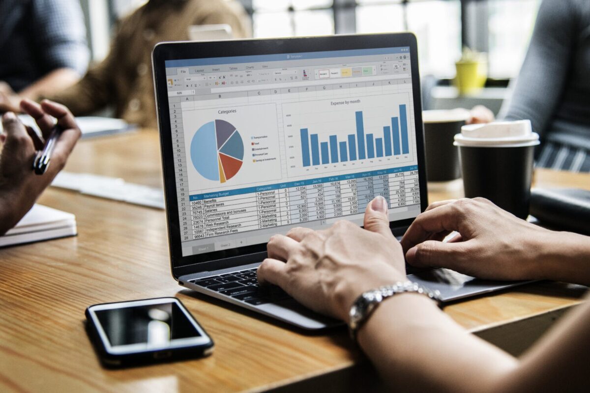 A person is typing on a laptop displaying business reports, including a pie chart and bar graphs, during a meeting.