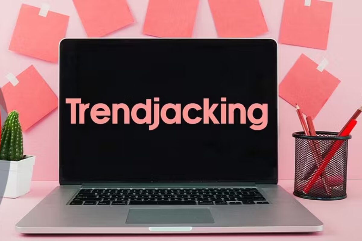 A laptop screen displaying the word "Trendjacking" in large red letters on a black background, set up on a pink desk with sticky notes and office supplies.