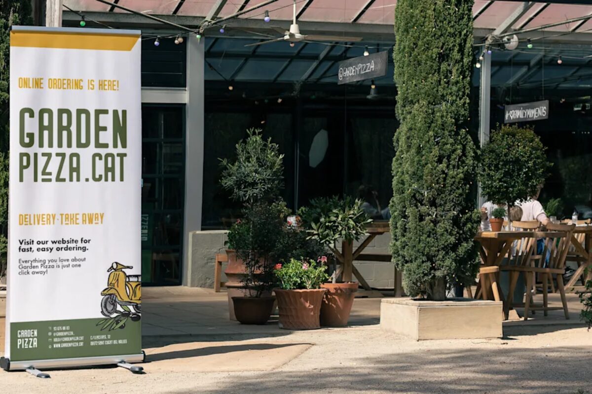 A banner advertising online ordering for a restaurant named Garden Pizza, shown outdoors in a shaded patio dining area with wooden tables, chairs, and potted plants.