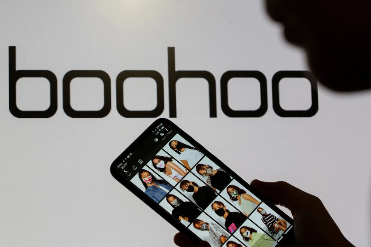 A hand holds a smartphone displaying a grid of fashion models on the Boohoo website, in front of a large, backlit Boohoo logo.