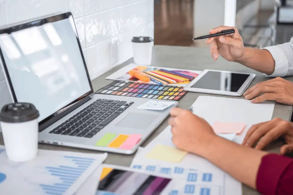 Two people working together at a desk on a laptop and tablet with color swatches and business documents.