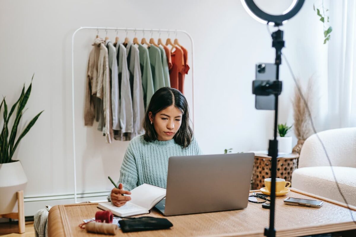 A woman working on a laptop and writing in a notebook at a desk in a room that looks like a small business or home office, with a clothing rack and a ring light visible