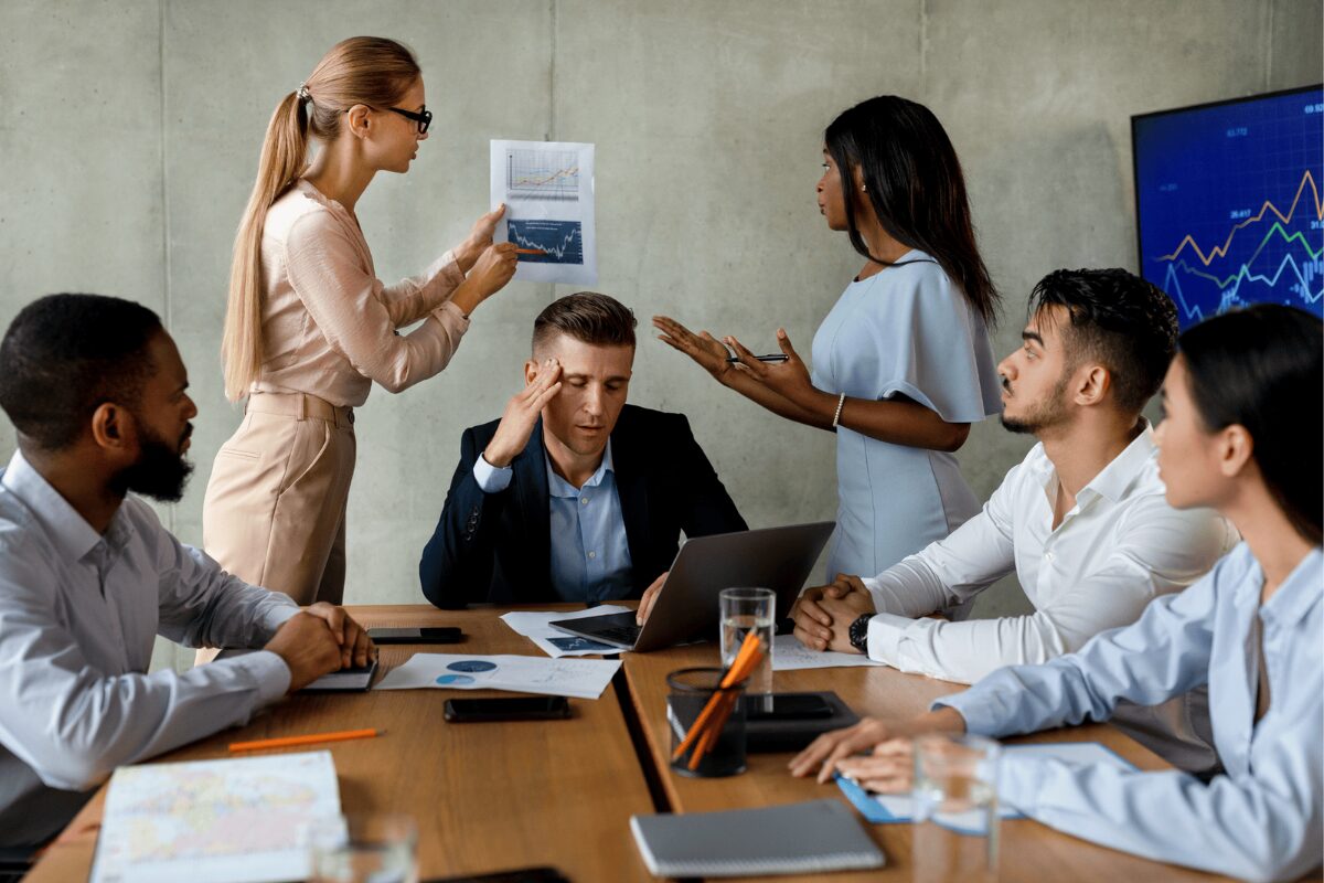 A tense business meeting where one woman presents a chart while another woman argues a point to the group, surrounding a stressed man at a table with a laptop and documents.