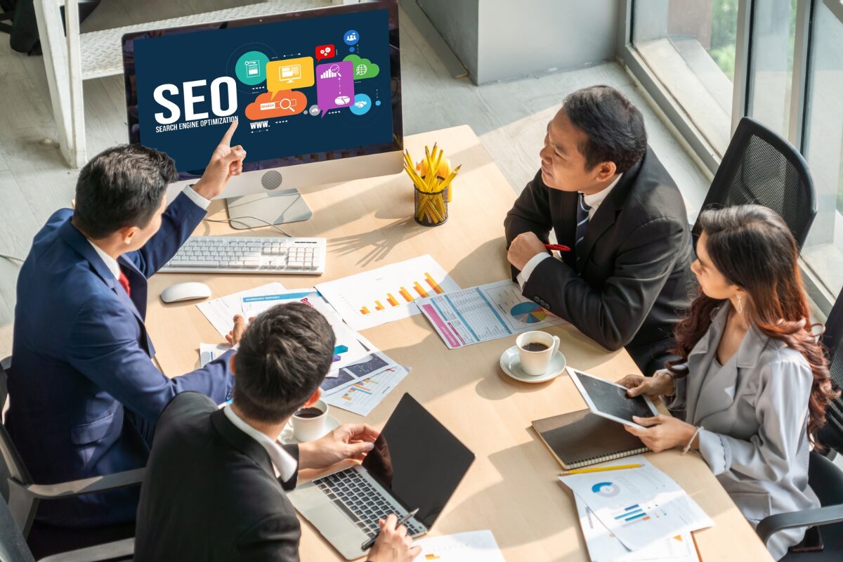 A group of four business people is holding a meeting and looking at a computer monitor displaying an illustration for Search Engine Optimization (SEO) concepts and icons.