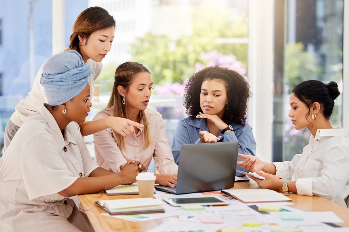 Five professional women collaborating around a conference table with a laptop and documents.