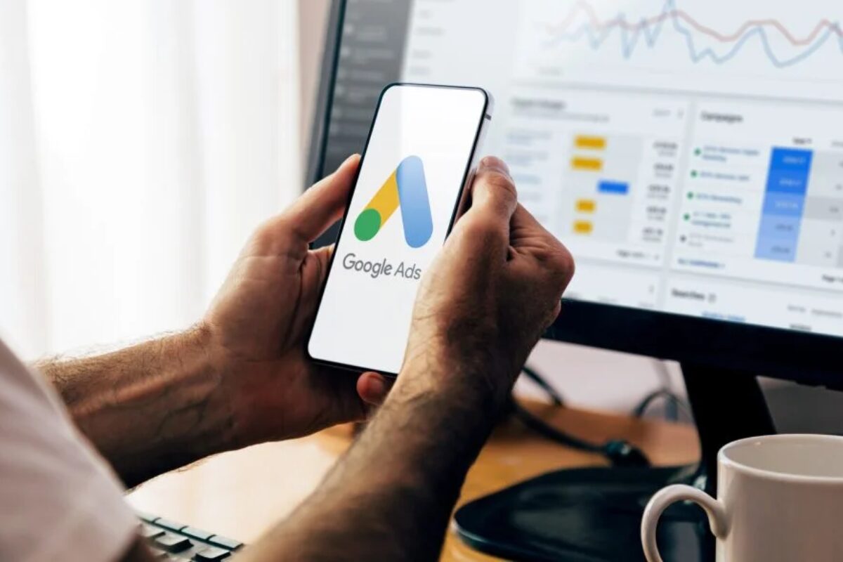 A person holds a smartphone displaying the Google Ads logo, with a computer monitor showing a marketing dashboard in the background.
