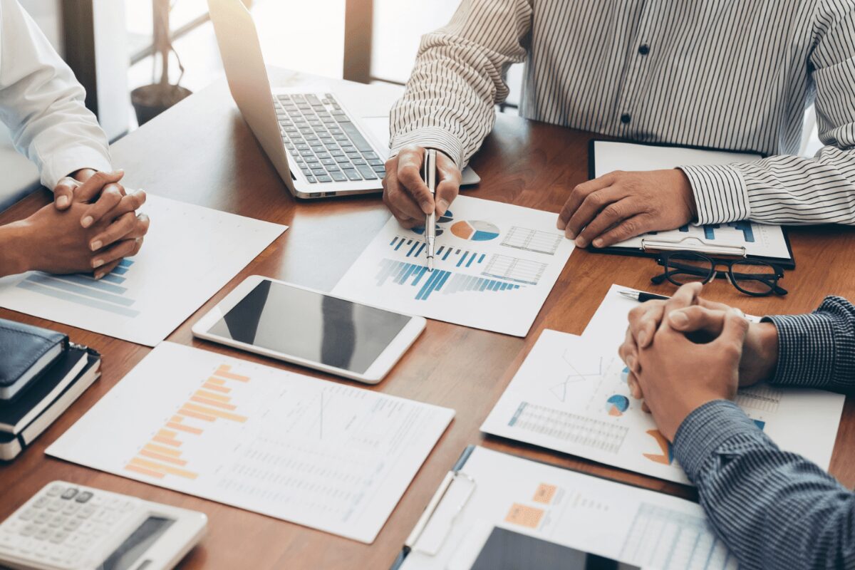 A collaborative business meeting taking place around a wooden conference table with participants reviewing financial documents, a laptop, and a tablet.
