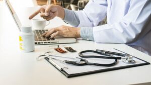 A medical professional in a white lab coat points at a laptop screen while typing with the other hand. A white pill bottle, blister packs of medication, a syringe, a stethoscope, and a clipboard are organized on the white desk in the foreground.