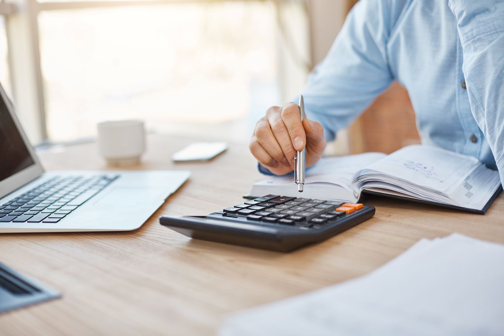 A close-up of a person in a blue shirt sitting at a wooden desk, using a silver pen to press buttons on a black calculator next to an open notebook and a laptop.